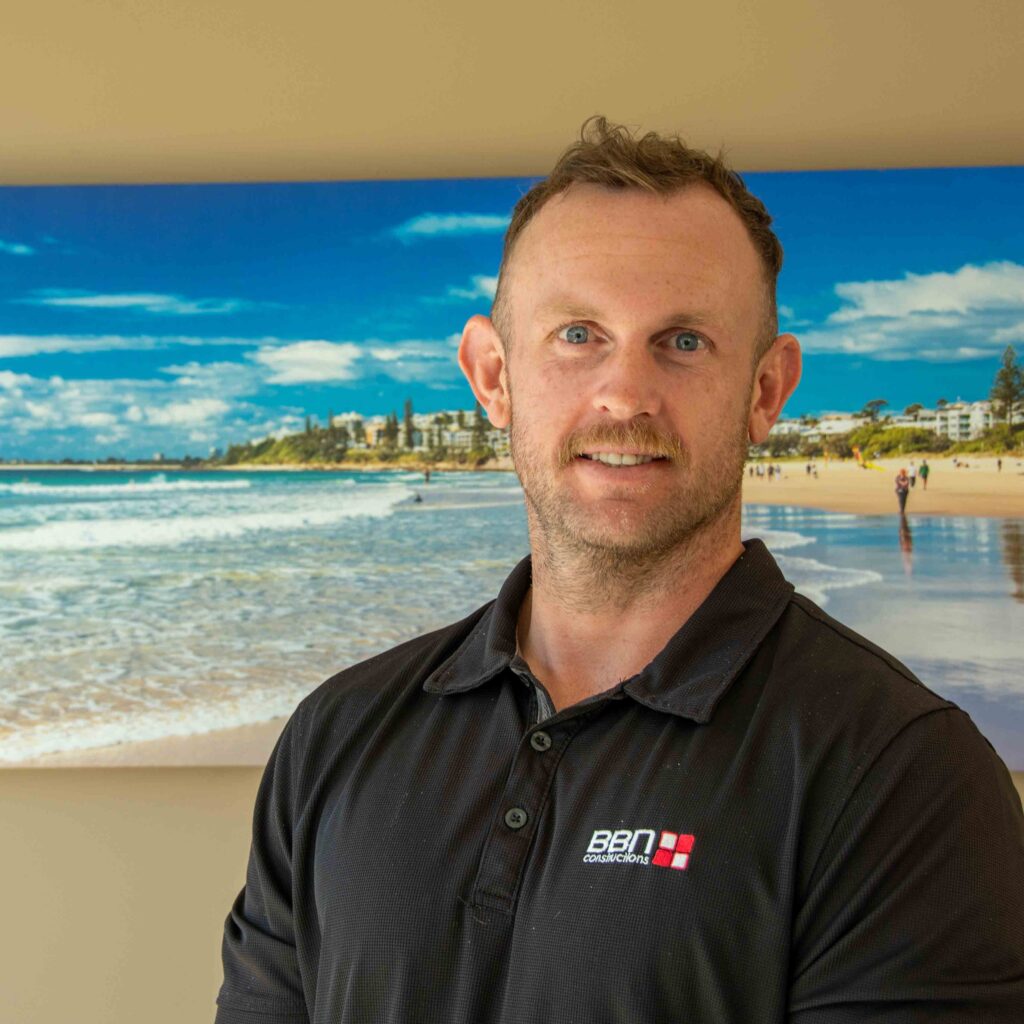 Portrait of Joel, a team member at BBN Constructions, wearing a black company polo in front of a beach-themed artwork.