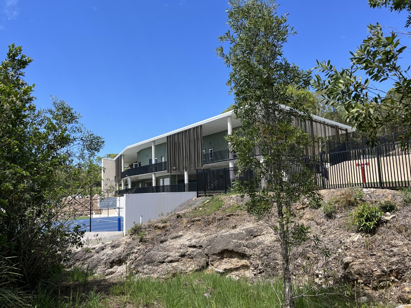 Exterior view of Coolum Beach Christian College building on a hillside with trees and a blue sports court under a clear sky.