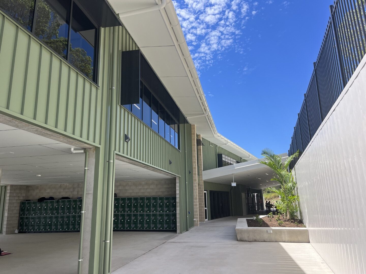 Covered walkway at Coolum Beach Christian College with green building facade, lockers, and corrugated fence under a blue sky.