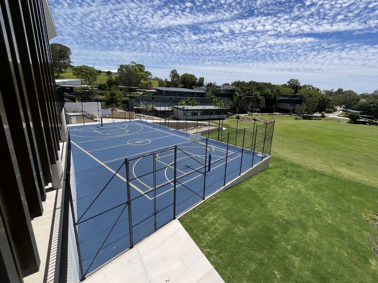 Aerial view of an outdoor blue sports court at Coolum Beach Christian College, surrounded by a fence, with grassy field and school buildings in the background.